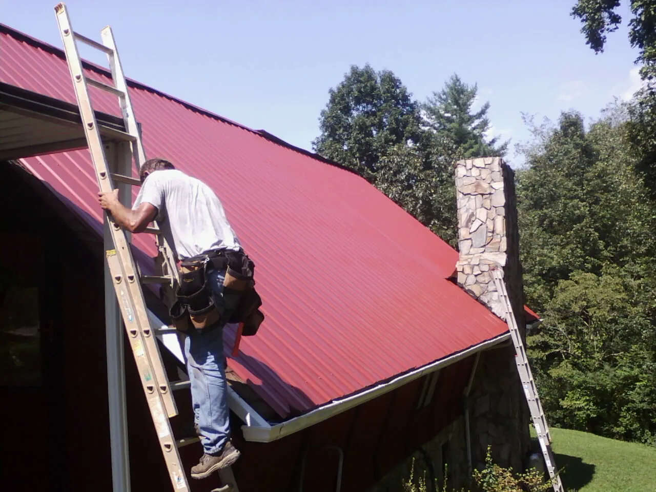 Roofer on a ladder beside a red metal roof and stone chimney — Soffit & Fascia Repair by Roofing Brothers in Harrison Township
